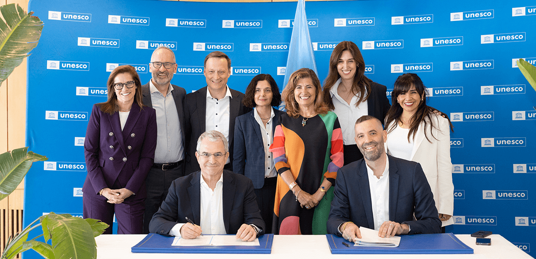 A group of professionals stands behind two individuals signing documents, with a UNESCO backdrop.
