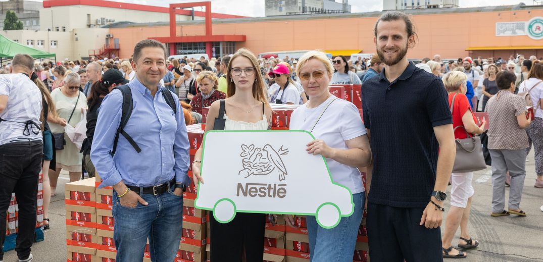A group holds a Nestlé logo sign amidst a large crowd, with stacks of boxes behind them at an outdoor event.
