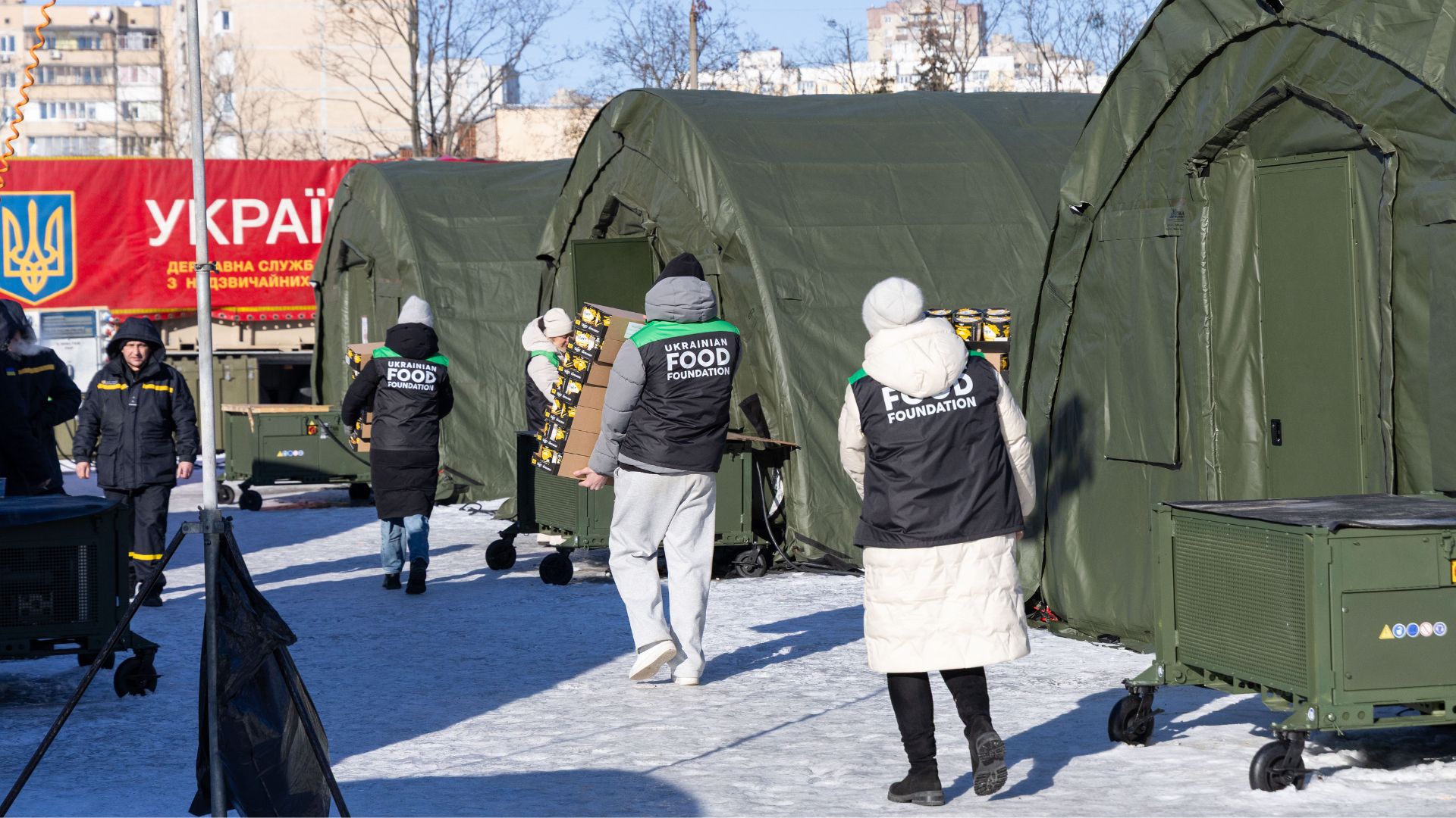 Volunteers from the Ukrainian Food Foundation carrying boxes near green tents in a snowy outdoor relief distribution area.