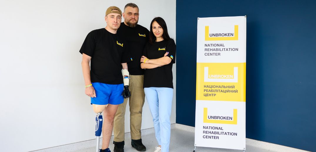 Троє осіб у однакових чорних футболках впевнено стоять біля банера Національного реабілітаційного центру «Unbroken»
