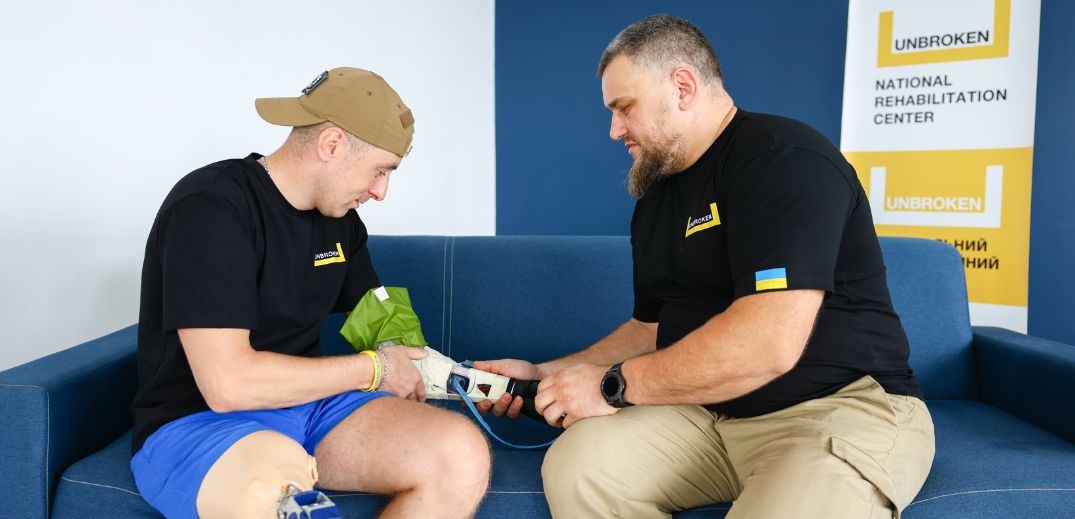 Двоє чоловіків взаємодіють на синьому дивані в реабілітаційному центрі, один використовує медичний пристрій на руці іншого.