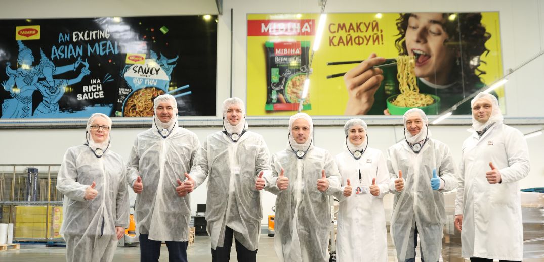 Group of eight people in protective white coats and masks standing indoors, giving thumbs up, with colorful product advertise