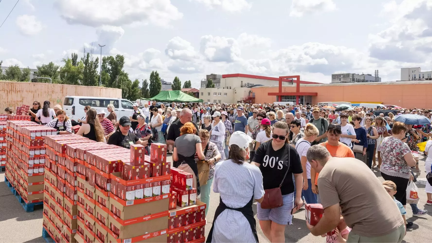 A crowded outdoor event with long lines, stacked boxes of products, and a mix of people socializing under a sunny sky with clouds